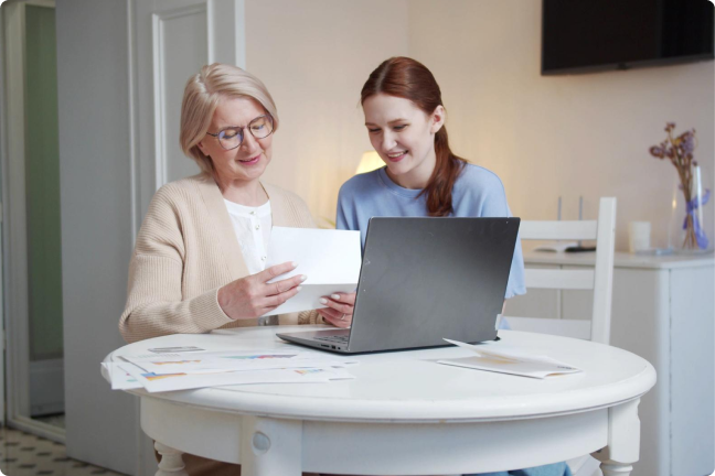An old and young woman holding a paper while smiling, and a laptop on top of a white round table