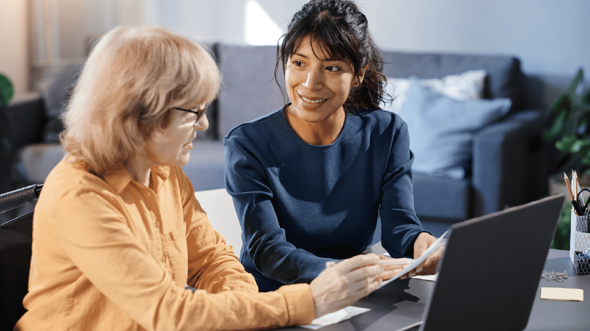 Two people sit at a desk, the one holding a paper while discussing business, a laptop on the top of table