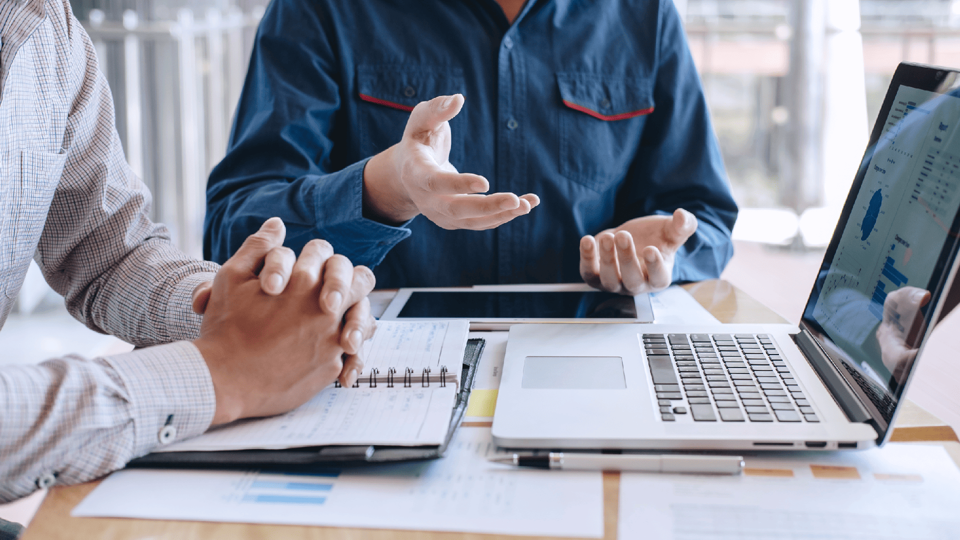 Two people sit at a desk reviewing documents, charts, and a laptop with data on the screen. One person gestures with their hands while the other listens with hands clasped, suggesting a business discussion or meeting.