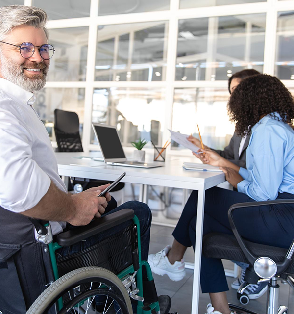 An old man smiling while sitting in a wheelchair, two ladies on the side talking, and a laptop on the table