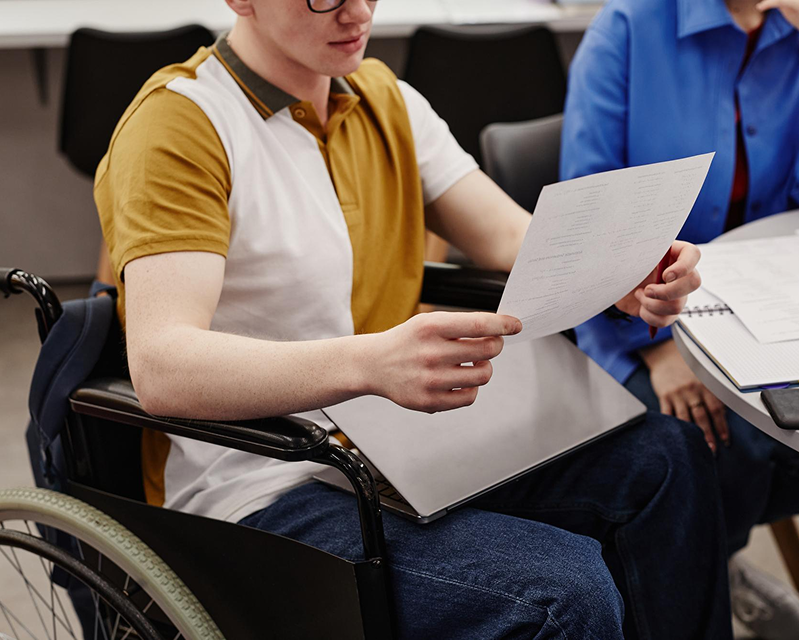 A man sits in a wheelchair wearing eyeglasses while holding a paper, and a man on his side wearing blue long sleeved